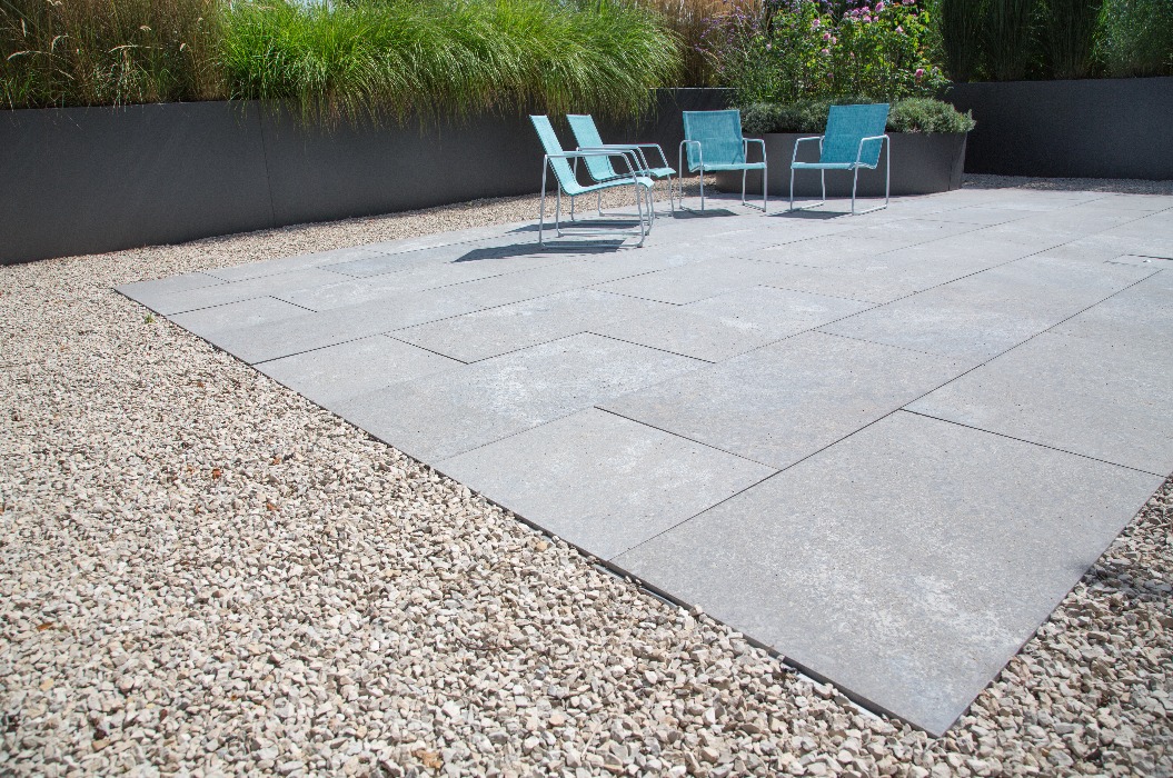 Moderne Garten- und Terrassengestaltung im Materialmix: Terrasse aus Steinplatten umgeben von Schotter und Metall Pflanzgefäßen mit Grünpflanzen